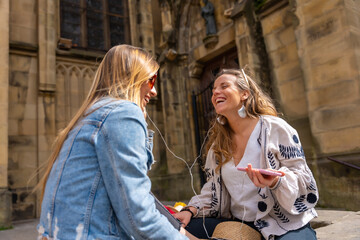 Listening to music in the city, two Caucasian female friends smiling and sharing headphones and...