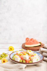 Vegetarian salad with watermelon, feta cheese, and grapes on blue ceramic plate on white wooden background. Side view, selective focus, copy space.