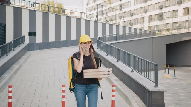 Caucasian young female courier calling a customer on the phone. A courier with a bag brings boxes to customers outdoors. The courier talks on the phone and walks down the street.