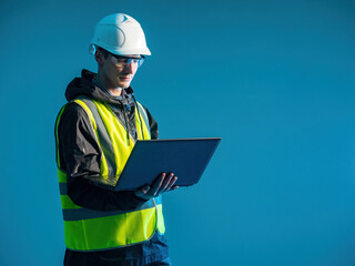 Worker stands with laptop in his hands. Computer master in working uniform. Computer engineer on blue. Computer master in yellow vest and hard hat. Engineer is working on laptop while standing.