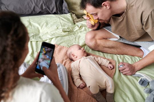 Young woman with smartphone taking photo of her husband playing with their adorable baby son on double bed at leisure