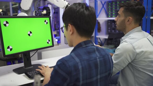 Back side view of Asian male data engineer sitting at desk keying a software code on a mock-up green screen, with Caucasian male colleague sitting testing the work of a robotic arm machine next to him