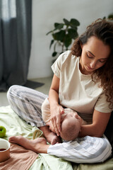 Young brunette woman looking at sleeping baby on her hands while sitting on double bed in the morning and having breakfast