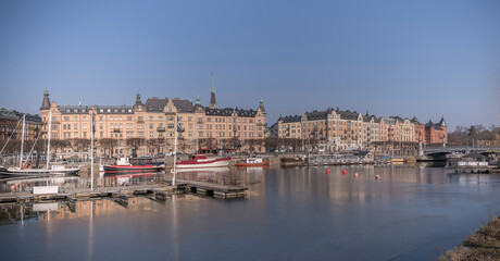 Obraz na płótnie Canvas Panorama view over the pier at the district Östermalm, boats and jetties in the icy water of the canal Djurgårdskanalen in the bay Nybroviken a sunny spring day in Stockholm