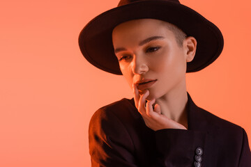 sensual woman with natural makeup, in black fedora hat, posing with hand near face isolated on coral.