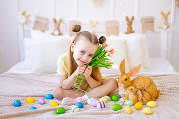 easter, a little girl with painted colored eggs and a rabbit holding spring flowers tulips at home in a bright room preparing for the holiday smiling and having fun