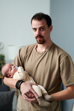 Young Man In Casual T-shirt Holding Baby Boy In Romper Suit While Standing In Front Of Camera And Lulling Infant