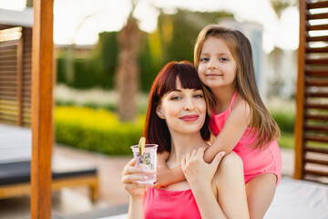 Portrait of a little daughter and her mother Drinking cold lemonade, joyfully smiling during a summer holiday break in a vacation villa on the sea. Daughter tenderly lovingly hugging her mother.