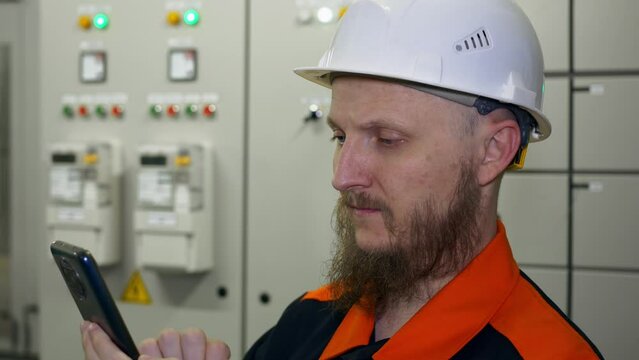 Engineer controls production equipment using smart phone in equipment control room in factory. A bearded man with blue eyes in overalls and a hard hat at work.