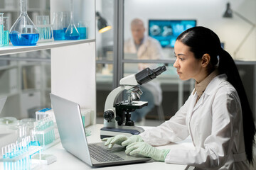 Young contemporary female chemist in whitecoat and gloves typing on laptop keyboard while looking at screen in laboratory - obrazy, fototapety, plakaty