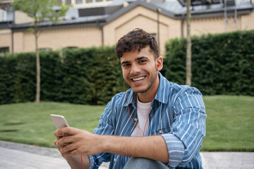 Smiling Indian hipster listening music outdoors looking at camera. Young handsome asian man using mobile phone, chatting, communication online