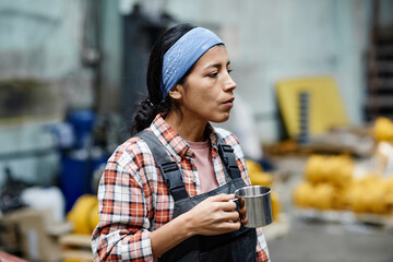 Young serious female builder in coveralls and blue headband holding mug with tea while having break in warehouse or workshop