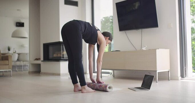 woman in sportswear doing stretching exercises while watching yoga training class on computer laptop online. Healthy girl exercising in living room with sofa couch in the background..