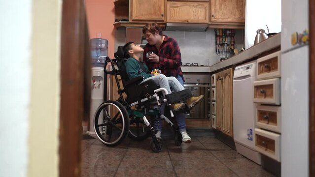 mother with disabled son in wheelchair cooks at home while giving him a glass of water - family with disabled child