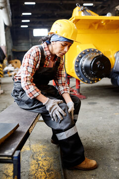 Young female engineer in protective helmet and coveralls sitting on bench and touching her injured knee after accident at work