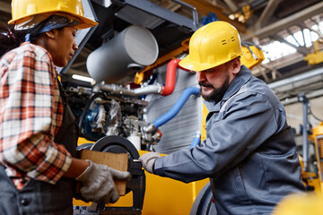 Mature workman in hardhat and workwear fixing shaft of industrial machine while his female...