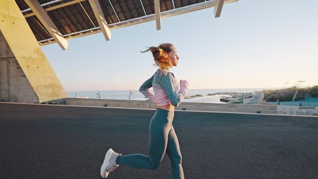 Tracking shot young woman with trained slender body enjoy jogging physical training outdoors move by running track on seafront in seaside city at morning or evening sunlight. Side view in slow motion