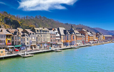 Fototapeta na wymiar Dinant, Belgium - March 9. 2022: View over river meuse on waterfront promenade with ancient colorful houses against blue winter sky, fluffy clouds