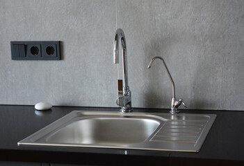 A metal kitchen sink on a black kitchen countertop with two water faucets, where one is a small...