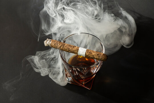 Whiskey Drinks With Cigars From Cuba Havana On Black Background. A Glass Of Whiskey Or Cognac With A Smoking Cigar Lying On Top. Top Down View.