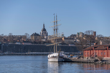Fototapeta na wymiar Sail boat, the brig Tre Kronor af Stockholm at a jetty on the island Kastellholmen and the district Södermalm with church and towers a sunny spring day in Stockholm