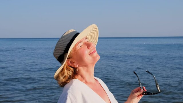 happy 50 year old woman in straw hat and sunglasses enjoying the sun against the blue sea background. Summer, vacation, vacation, active retirees