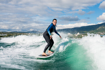 Young athletic female in wetsuit riding on endless waves behind a boat on sunny day. Woman practice wakeboarding, carving behind the baot. Watersport concept.