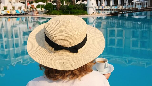 female in a straw hat wearing a white dress relaxing by the pool with a cup of coffee, vacation concept, good morning. woman sitting by the pool