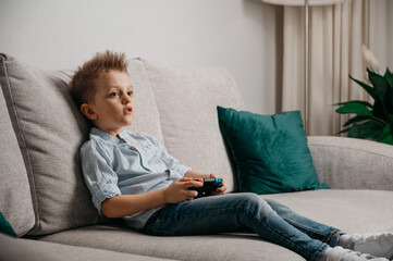 Happy boy playing video games holding game controller sitting on the coach in living room