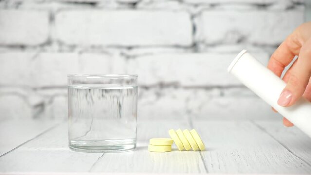 clear glass of water and tube of yellow soluble round effervescent tablets on white background. A woman's hand takes out liquid vitamins in close-up. Treatment and prevention of viral diseases.