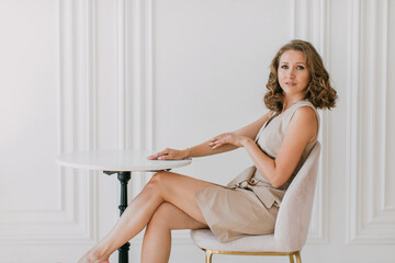 A young woman with curly hair, dressed in a light short formal dress, sits on a pink chair at a table against a white wall and smiles at the camera