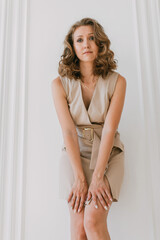 Bottom-up fashion portrait of young woman dressed in a short formal dress stands against a white wall with moldings and looks into the camera