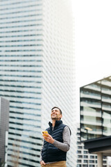 Freelancer male in casual clothes, using smartphone, modern building on background. Working remotely in urban location