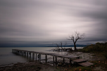 Fototapeta na wymiar View of a pier on a lake, beneath a dramatic, moody sky with skeletal, bare trees on the background