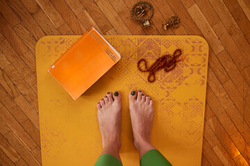 Top view of yogi woman's legs on the mat with book, bracelet and miniature.