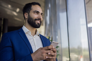 Handsome moroccan businessman holding smart phone shopping online looking at window standing in modern office. Middle easter male using mobile app, copy space. Successful business concept