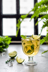 Iced tea in a beautiful glass with lime slices and thyme, stands on a table in backlight, a window on the back surface and flowers, a transparent tasty drink with vitamins