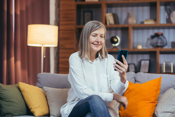 happy senior woman uses phone for online communication, sitting on sofa at home in the evening and smiling