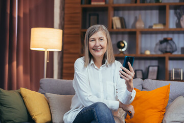 happy senior woman uses phone for online communication, sitting on sofa at home in the evening and smiling