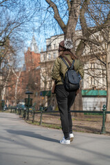 A lonely girl in a short jacket and sports trousers with a black bag behind her back walks along the asphalt in the city square on a sunny day in early spring. Back view