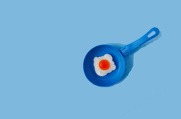 A blue frying pan with a fake scrambled egg on pastel blue background. Creative concept for Easter celebration card or banner. Surreal design for healthy breakfast