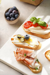 Italian antipasto bruschetta - white bread slices with smoked ham, goat cheese, blueberries, mozzarella, tomatoes, basil on marble board, glass with lemon water