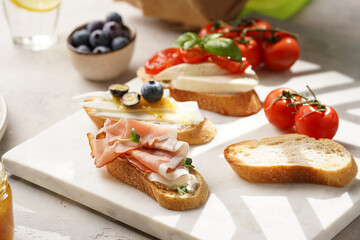 Italian antipasto bruschetta - white bread slices with smoked ham, goat cheese, blueberries, mozzarella, tomatoes, basil on marble board, glass with lemon water