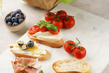 Italian antipasto bruschetta - white bread slices with smoked ham, goat cheese, blueberries, mozzarella, tomatoes, basil on marble board, glass with lemon water