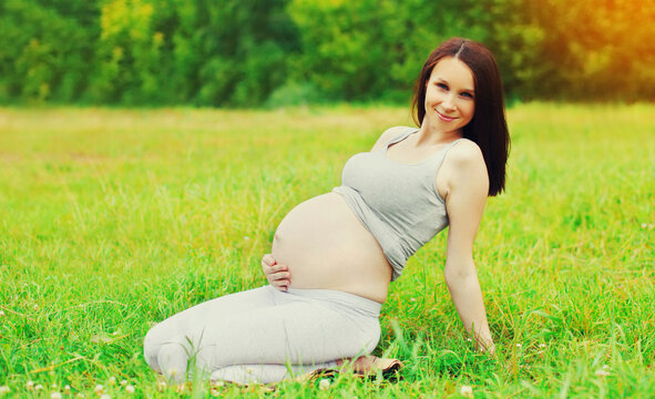 Portrait of happy smiling young pregnant woman in sunny summer park sitting on the grass