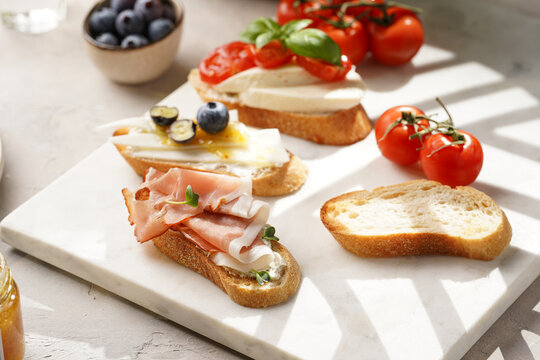 Italian antipasto bruschetta - white bread slices with smoked ham, goat cheese, blueberries, mozzarella, tomatoes, basil on marble board, glass with lemon water