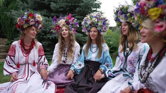 Positive Ukrainian young women singing sitting in national clothing in spring summer park. Group of confident beautiful ladies in traditional embroidered dresses and head wreathes outdoors