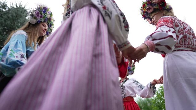 Bottom angle view of smiling adult woman standing outdoors as circle of young ladies in Ukrainian traditional dresses walking around holding hands. Happy lady enjoying folk dance in park. Slow motion