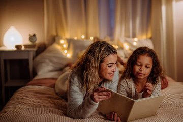 Happy mother with her little daughter lying on bed and reading book in evening at home.