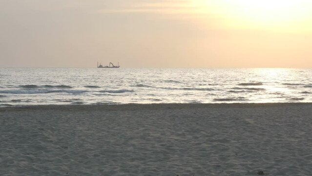 a dredge ship sails on the horizon, in a late afternoon warmed by the golden light of the sun
